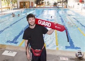 A life guard in a black shirt holding a red floater in front of a pool