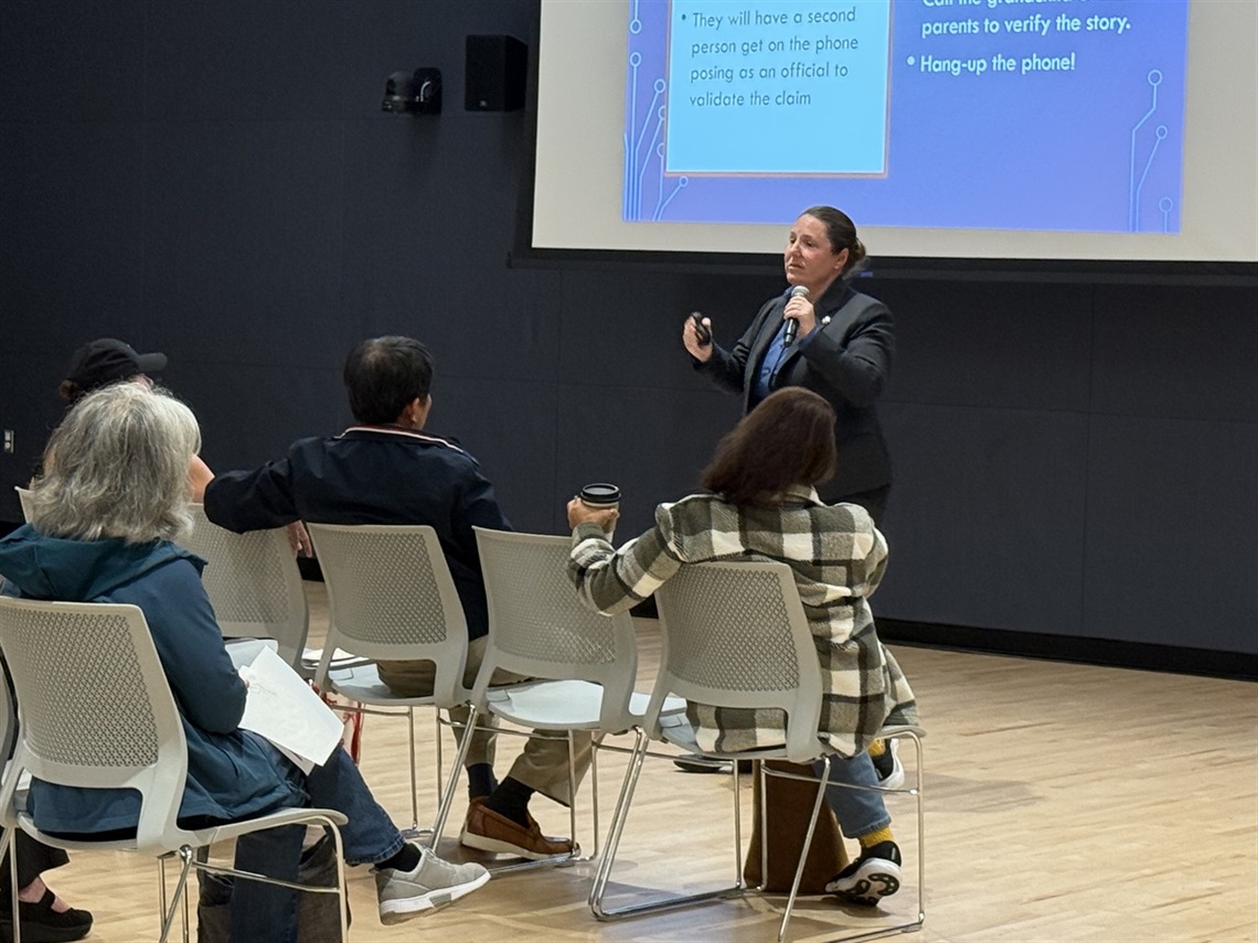 Detective Kathleen Walsh giving a seminar on how to identify scams in a light room with several people in the front row looking at a screen with the presentation