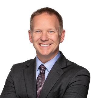 Drew Corbett's headshot. He is wearing a black suit with a light blue collared shirt and purple patterned necktie