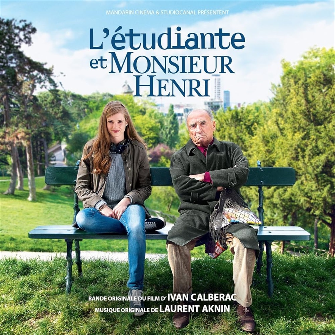 An older man with his arms crossed (Claude Brasseur) and a smiling young woman (Noemie Schmidt) sit on a park bench.