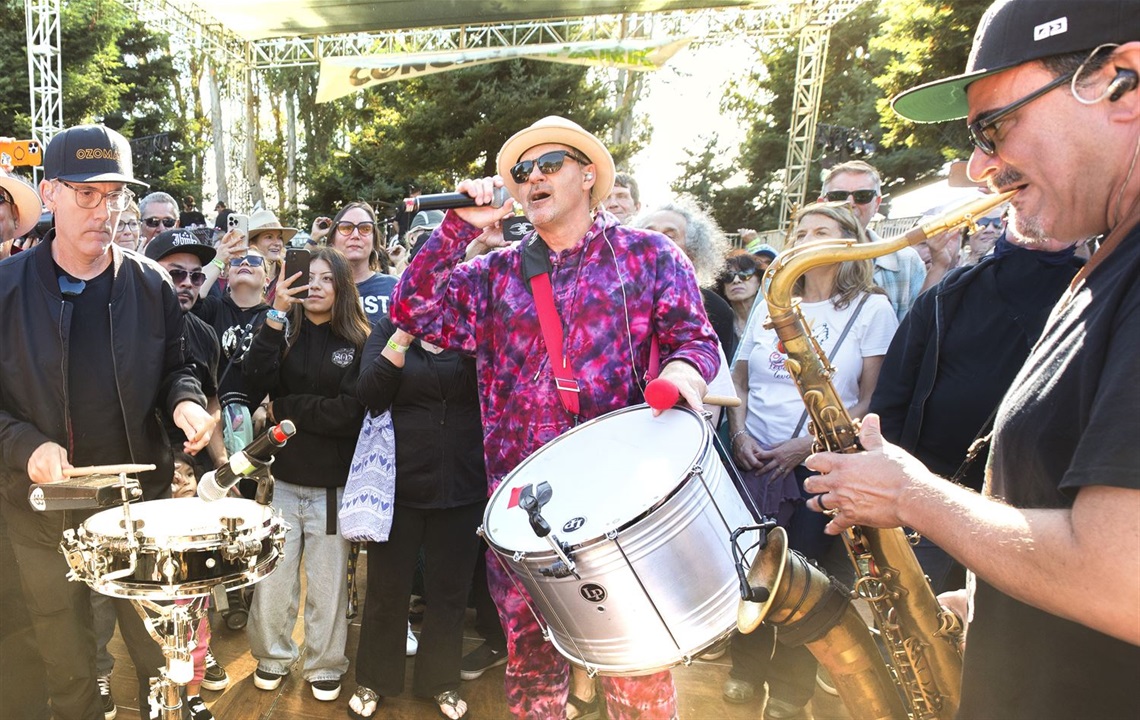 Outdoor concert event featuring live musicians performing among a crowd. A singer with a drum and band members on percussion and saxophone engage attendees in a lively, inclusive community setting.