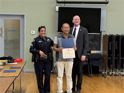 Senior male with gray vest, white shirt, and beige pants holding his graduation certificate next to Segeant Dominguez and Captain Plank .png