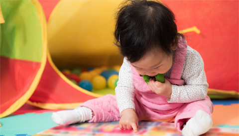 A baby wearing a white long sleeve and socks, pink overalls is sitting down and pointing at the play mat.