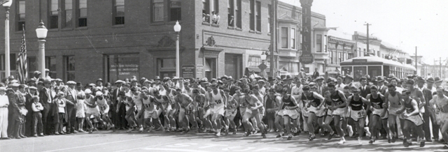 A historical photo in black and white of several people participating in a race on Grand Avenue with a trolley behind them.