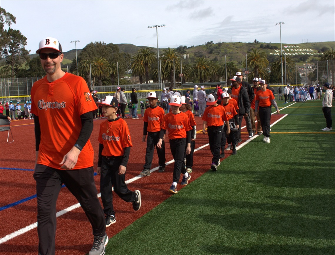 A youth baseball team in orange jerseys and black pants walks in a line during the  Parks & Recreation Youth Baseball Celebration. An adult coach leads, followed by children in matching caps, with teams, spectators, and green hills in the background.